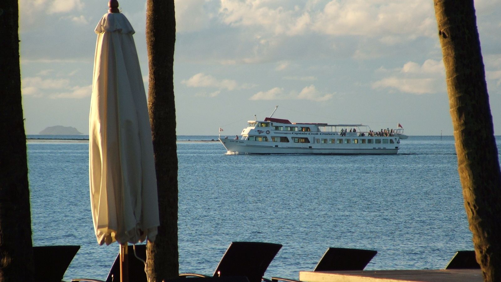 Wit passagiersschip vaart uit vanaf Denarau, gezien vanaf een strand met palmen en ligbedden